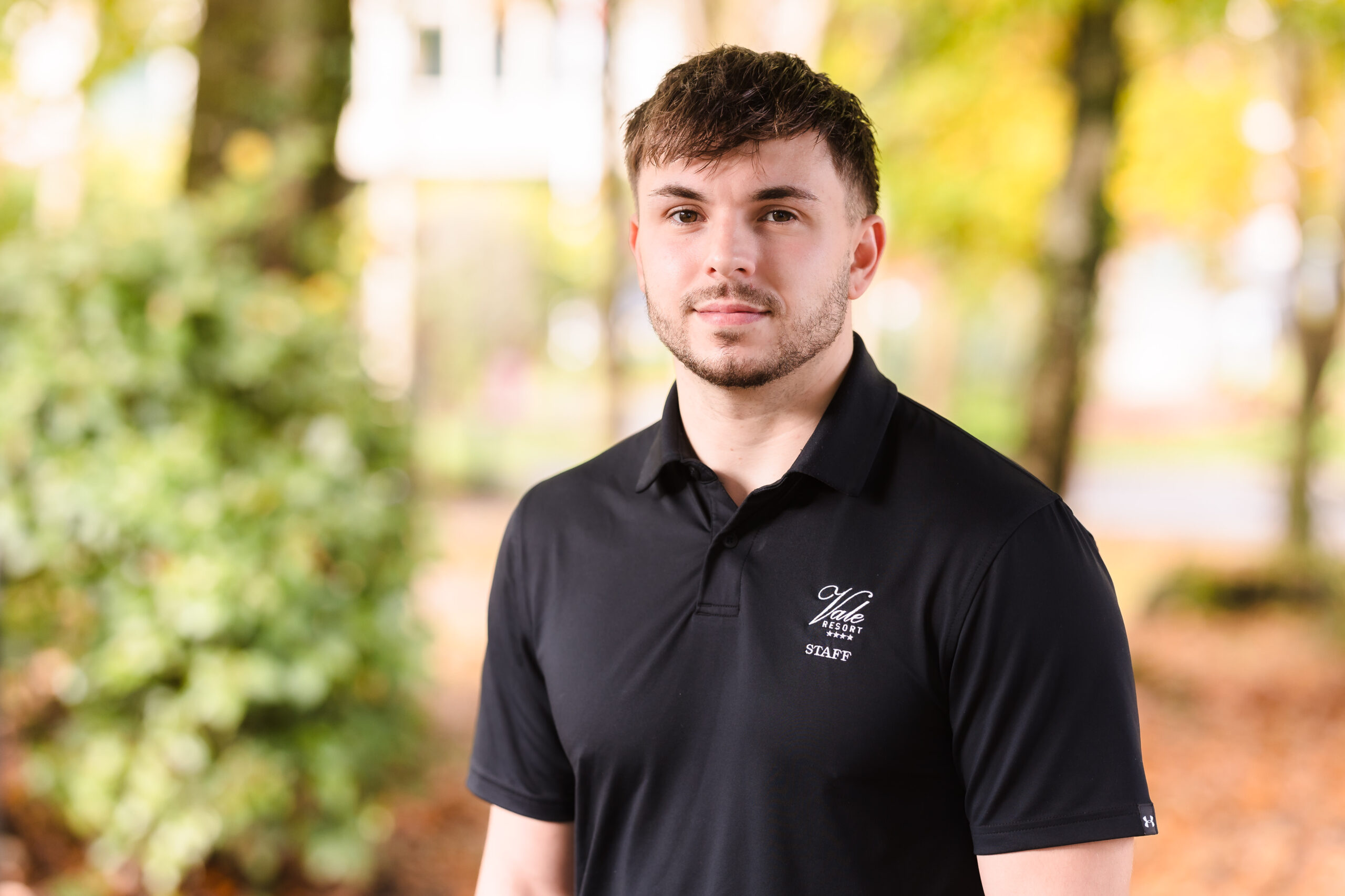 Man in a black polo shirt stands outdoors, with blurred greenery and trees in the background.