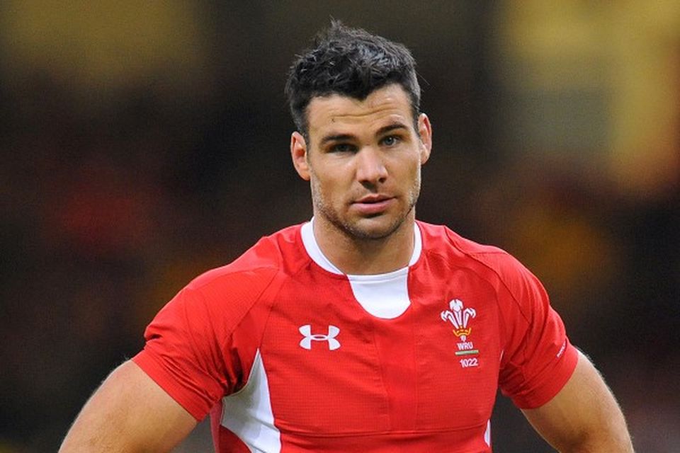 A rugby player in a red jersey with a white logo stands on the field, looking forward.