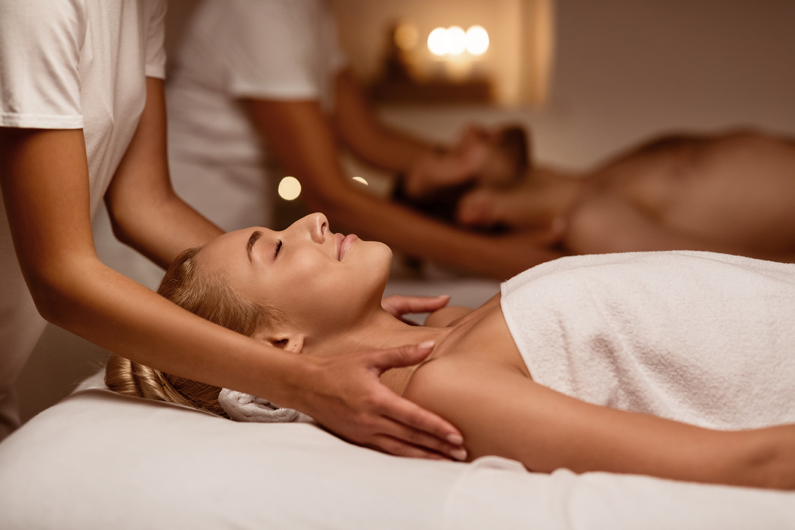 Two people receive massages in a spa, each lying on a massage table with towels covering them. Candles are lit in the background, creating a soothing atmosphere.