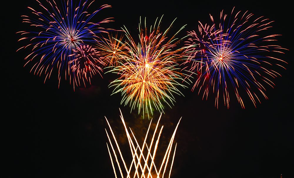 Colorful fireworks burst in the night sky, with vivid reds, blues, greens, and yellows creating dazzling patterns.