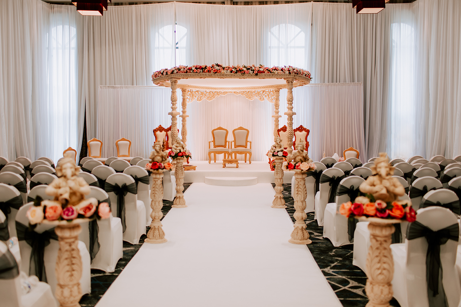 Ornate wedding mandap with floral decorations and two chairs, facing an aisle lined with flower arrangements and white-covered chairs.