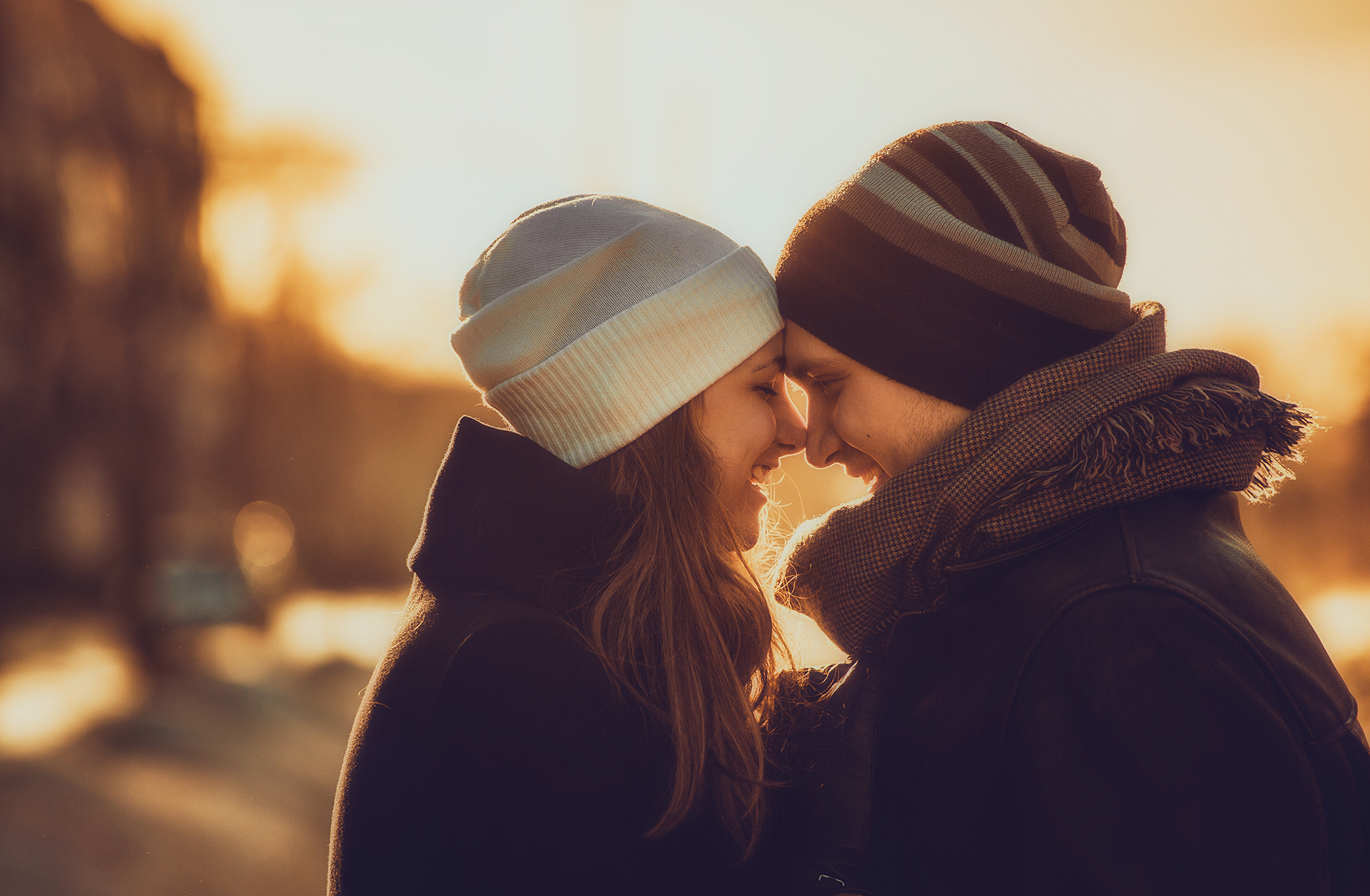 A couple in winter clothing touch noses and smile at each other during sunset.