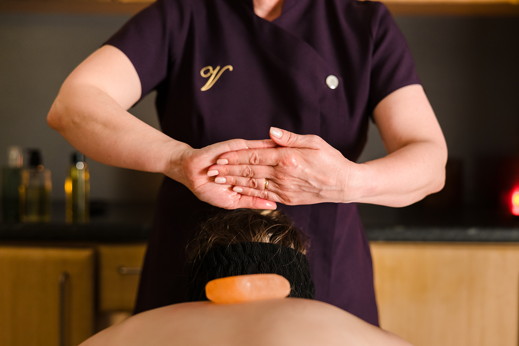 Massage therapist in a purple uniform performing a treatment on a person lying face down with a warm stone on their back.