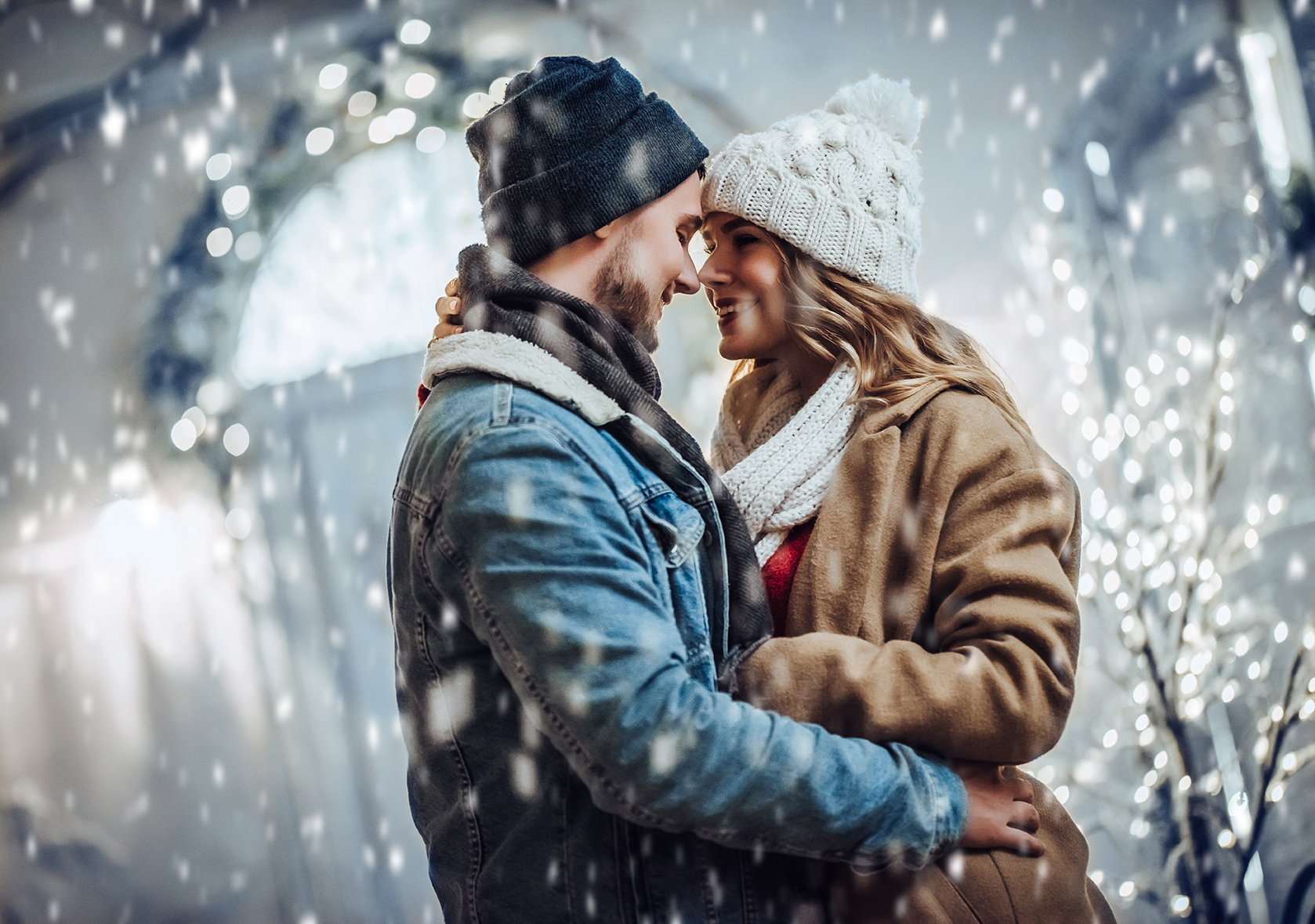 A couple embracing in snowy weather, both wearing winter coats and knitted hats. Snowflakes fall around them, with blurred lights in the background.