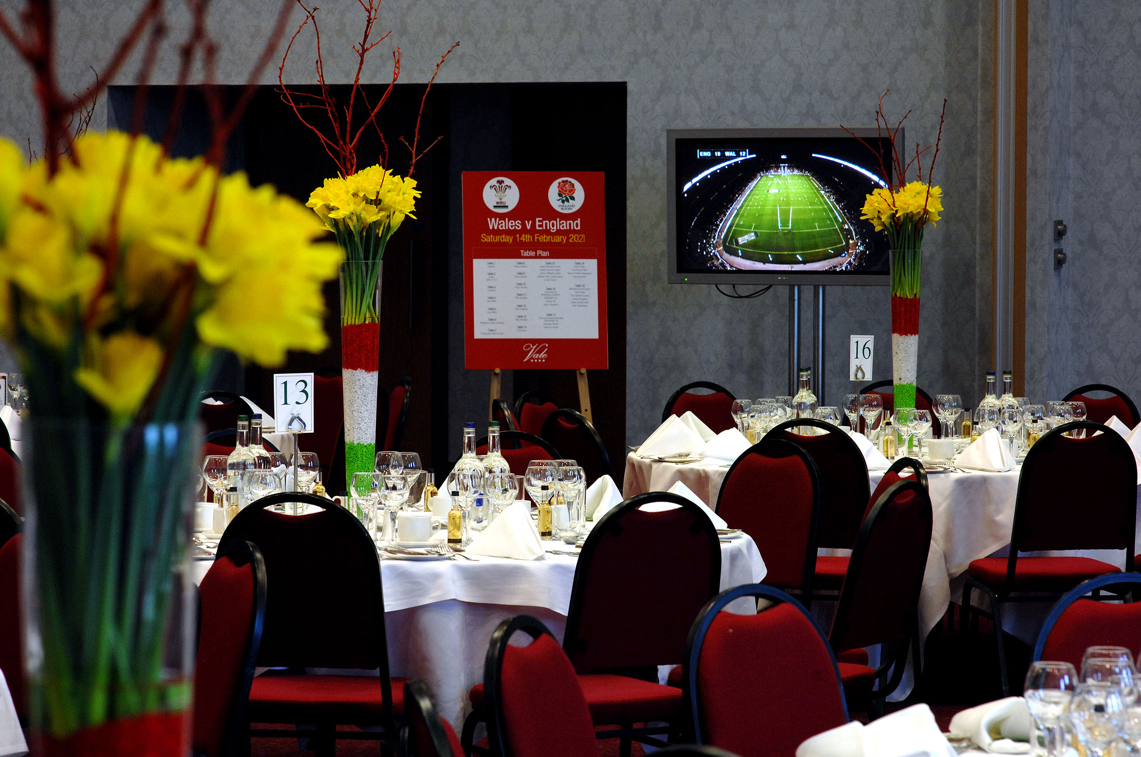 A banquet hall set for an event with round tables, yellow floral centerpieces, a display showing a rugby match, and a sign for a Wales vs. England game.