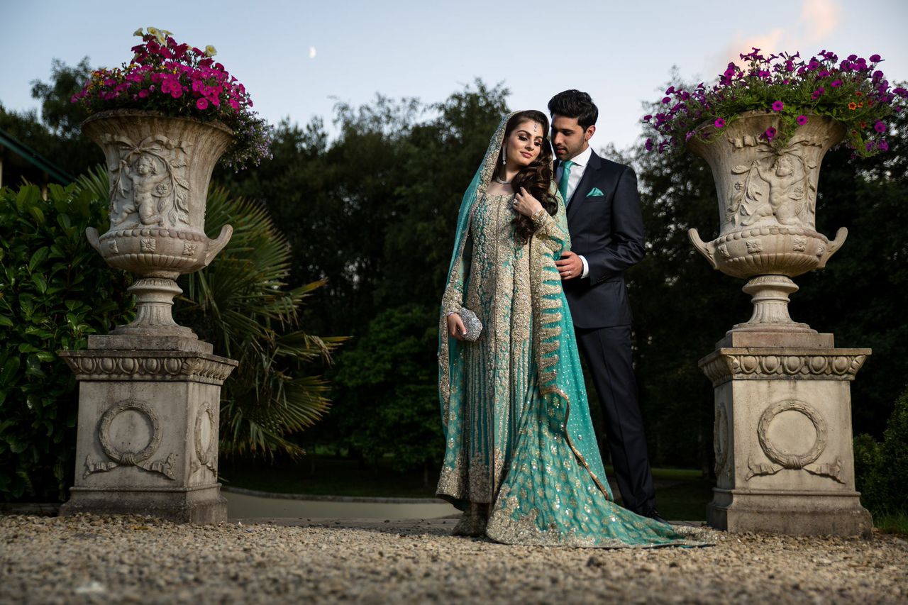 A couple in elegant attire poses outdoors between ornate flower urns against a backdrop of trees and a twilight sky.