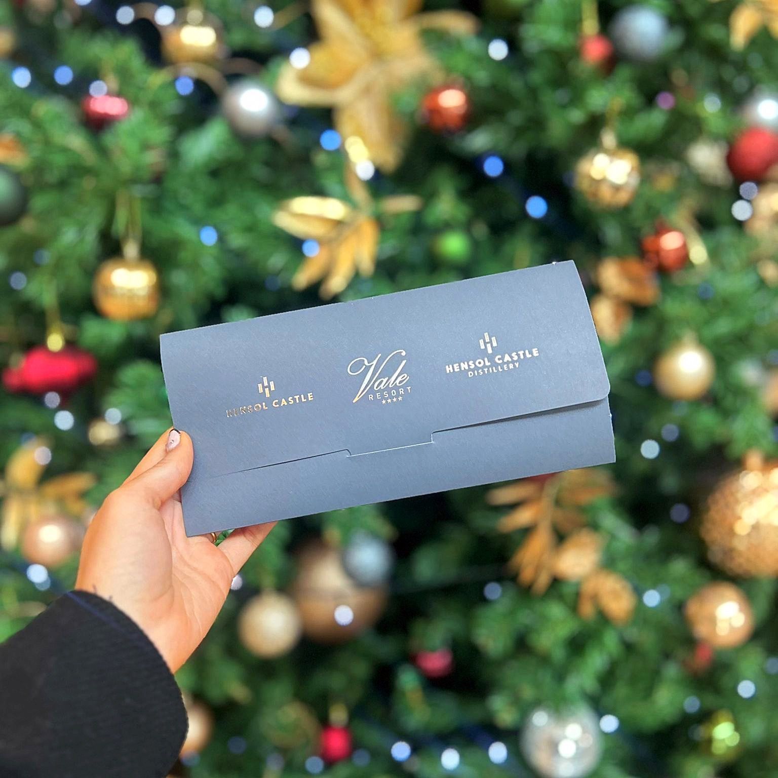 Hand holding a blue envelope in front of a decorated Christmas tree with ornaments and lights.