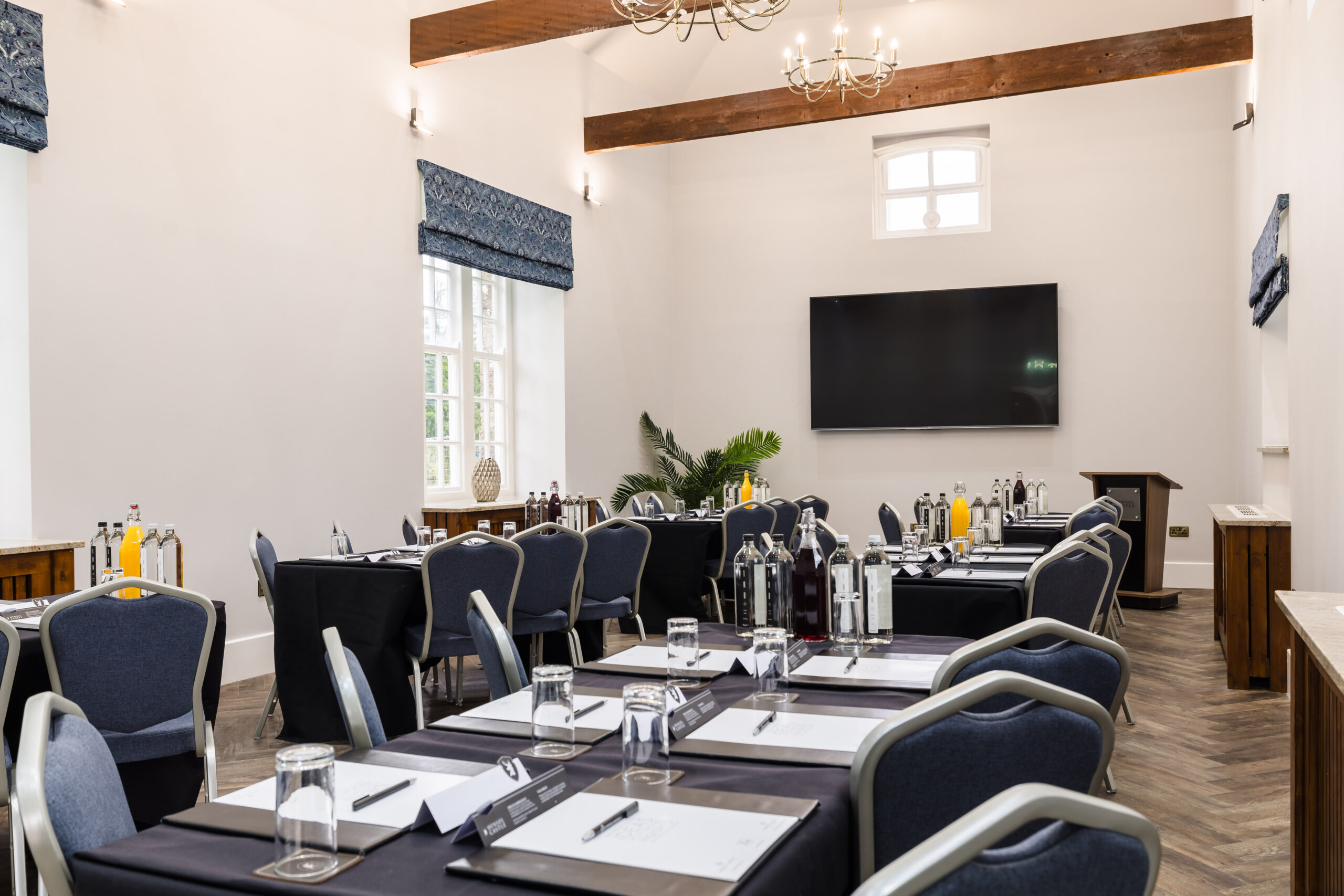 Conference room setup with tables and chairs, lined with bottled drinks, notepads, and glasses, facing a large screen.