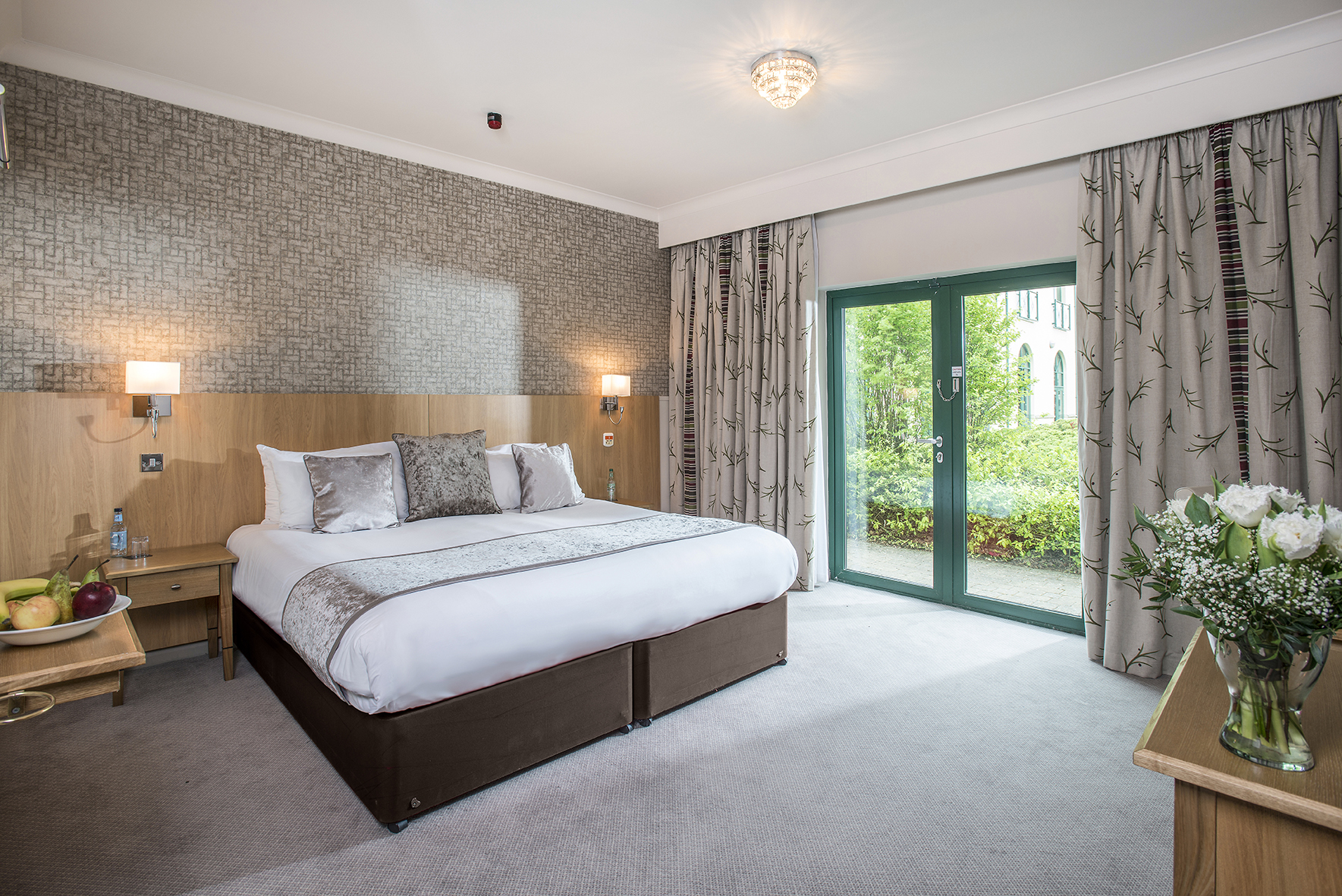 Hotel room with a king-sized bed, nightstands, decorative pillows, curtains, and a sliding glass door leading to a garden view.