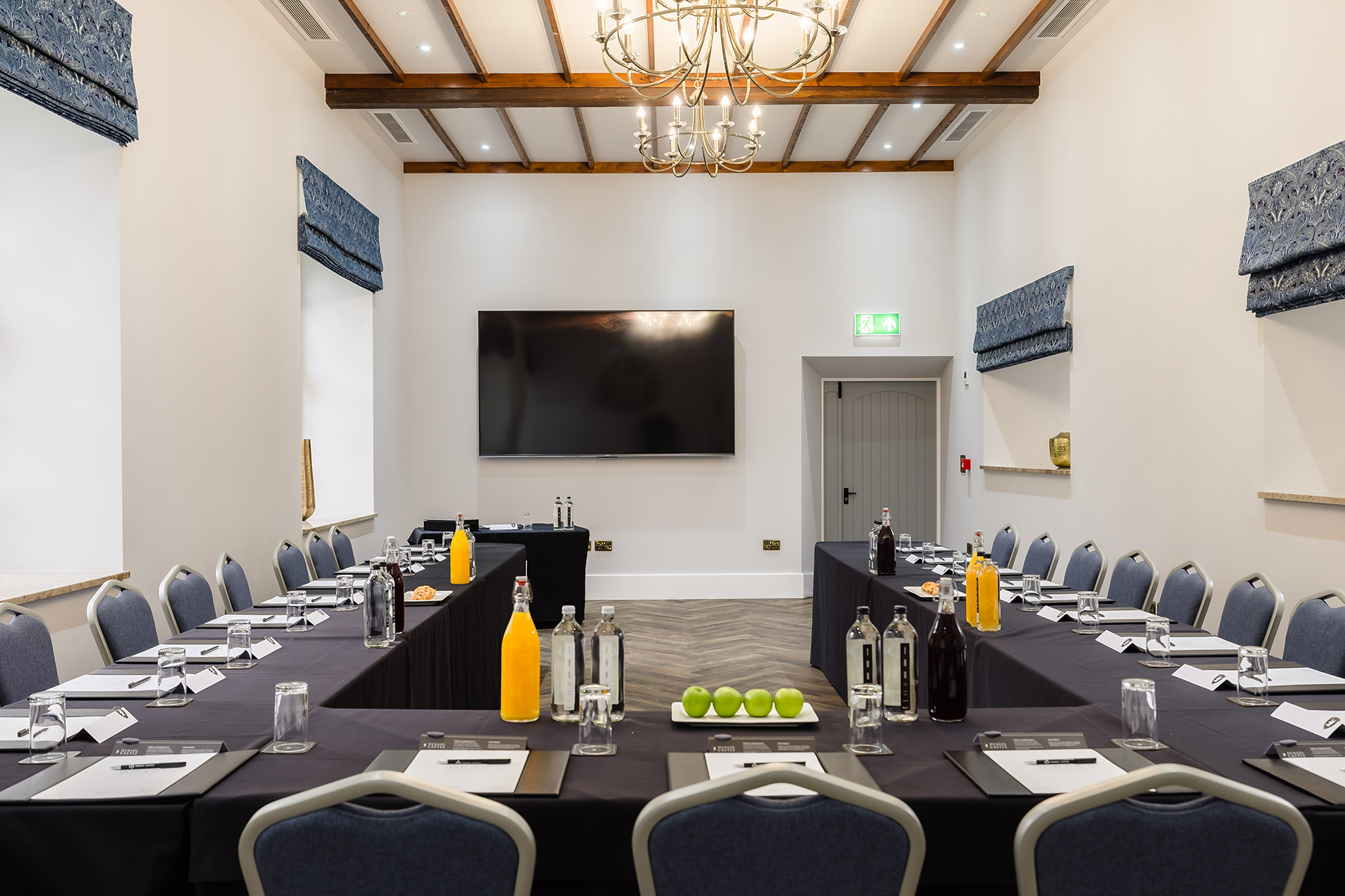 A conference room with a U-shaped table setup, notepads, water bottles, juice, and a bowl of green apples. A large screen at the front.