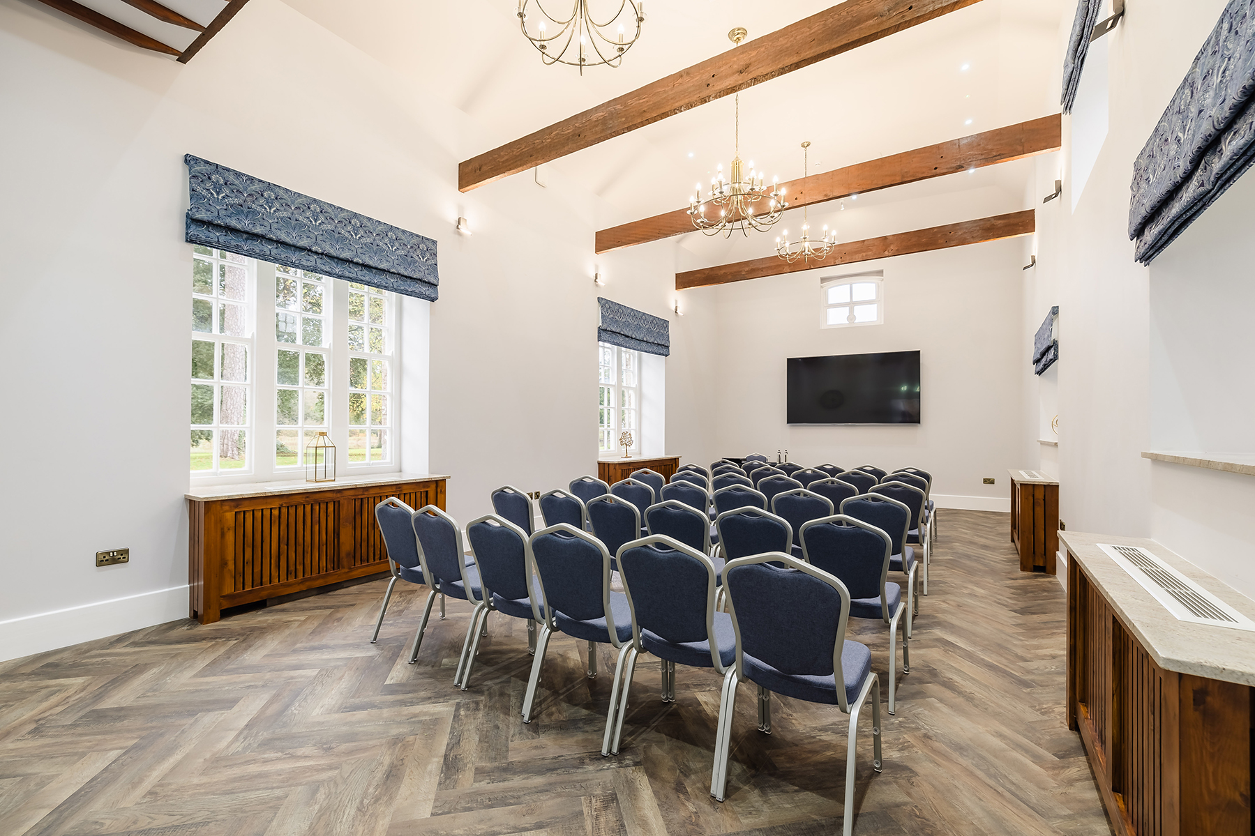 A well-lit conference room with rows of blue chairs facing a large wall-mounted screen, wooden beams, and chandeliers.