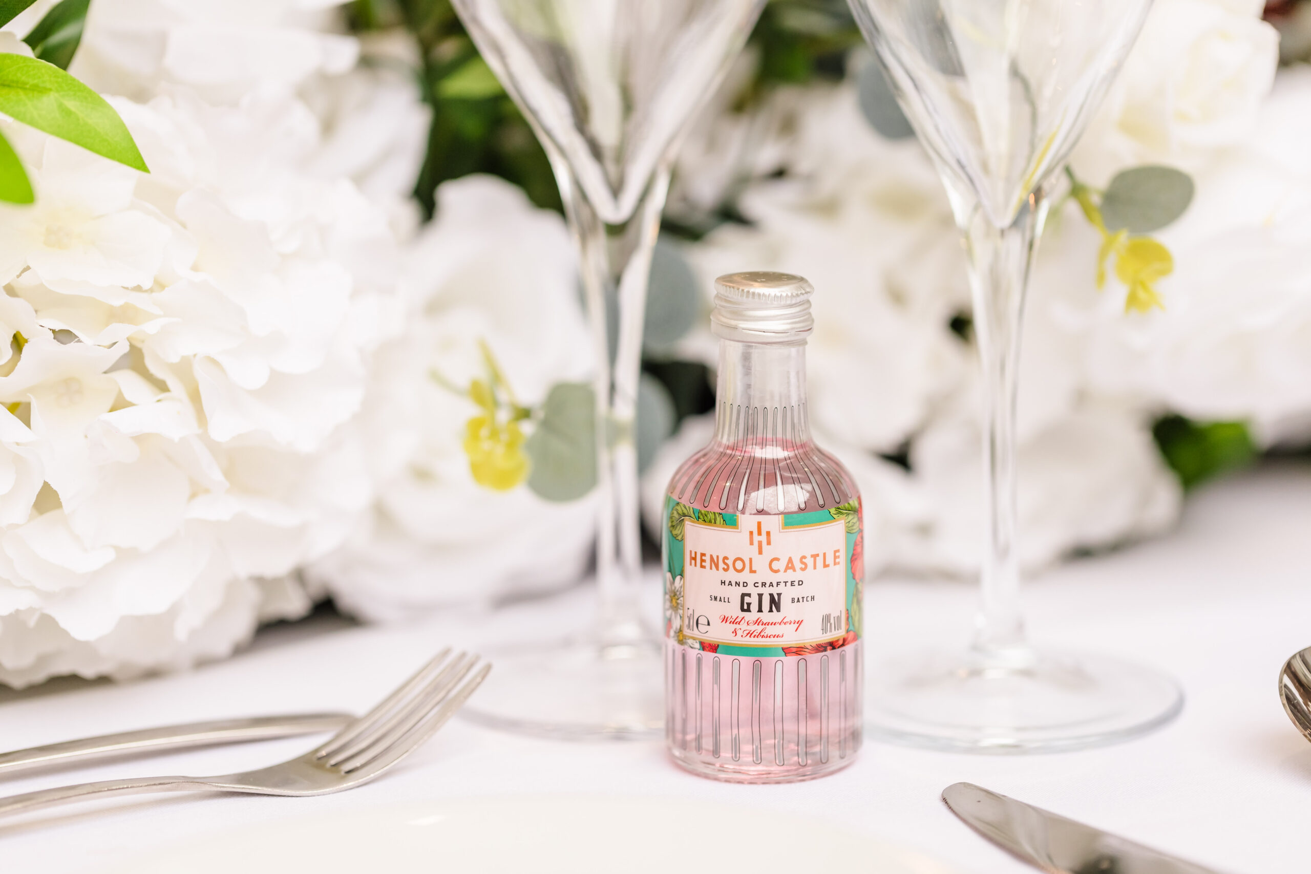 A small pink bottle of Hensol Castle gin on a table with two empty glasses and white flowers in the background.