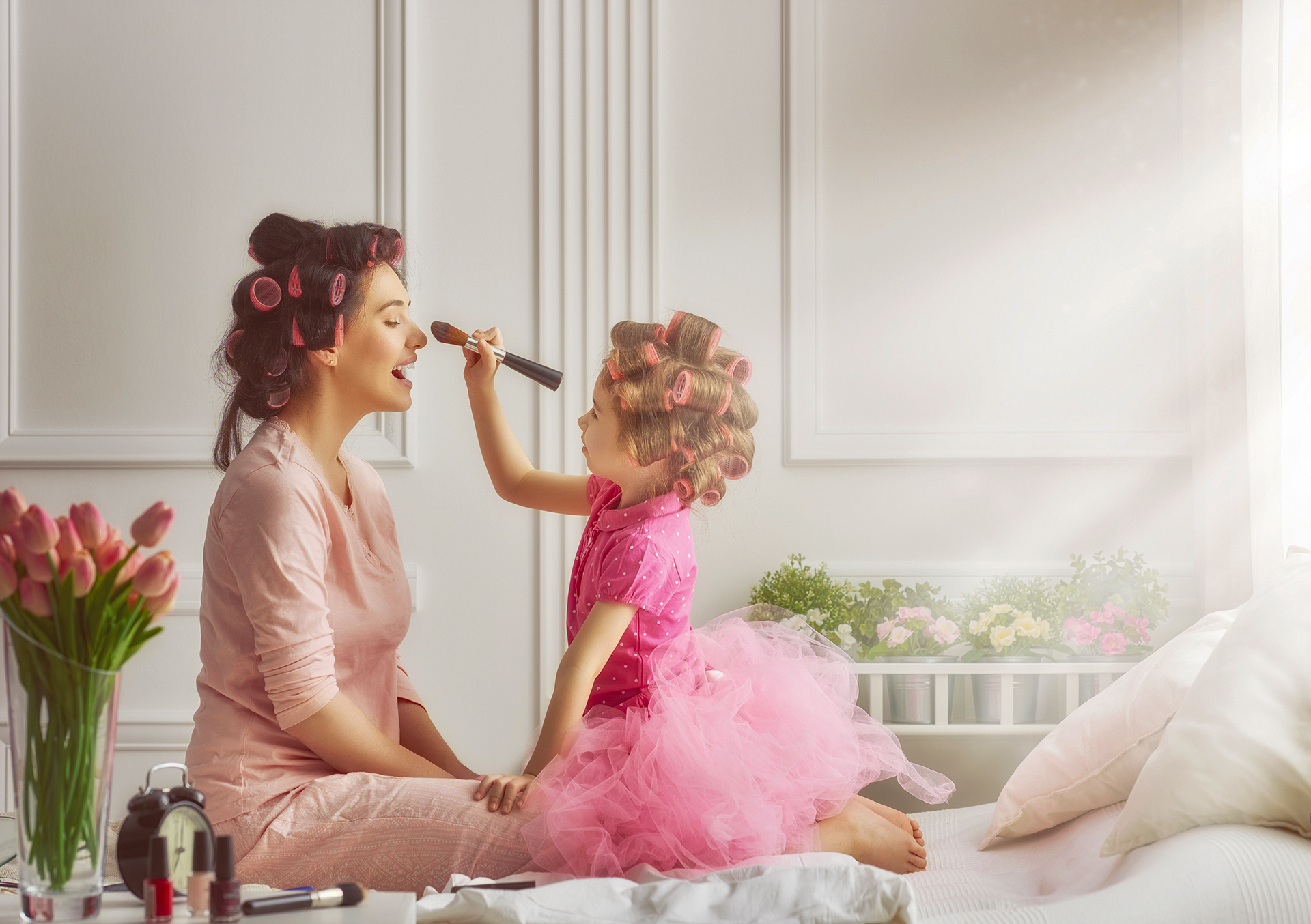 A woman and a young girl with hair rollers sit on a bed; the girl applies makeup to the woman’s face using a brush.