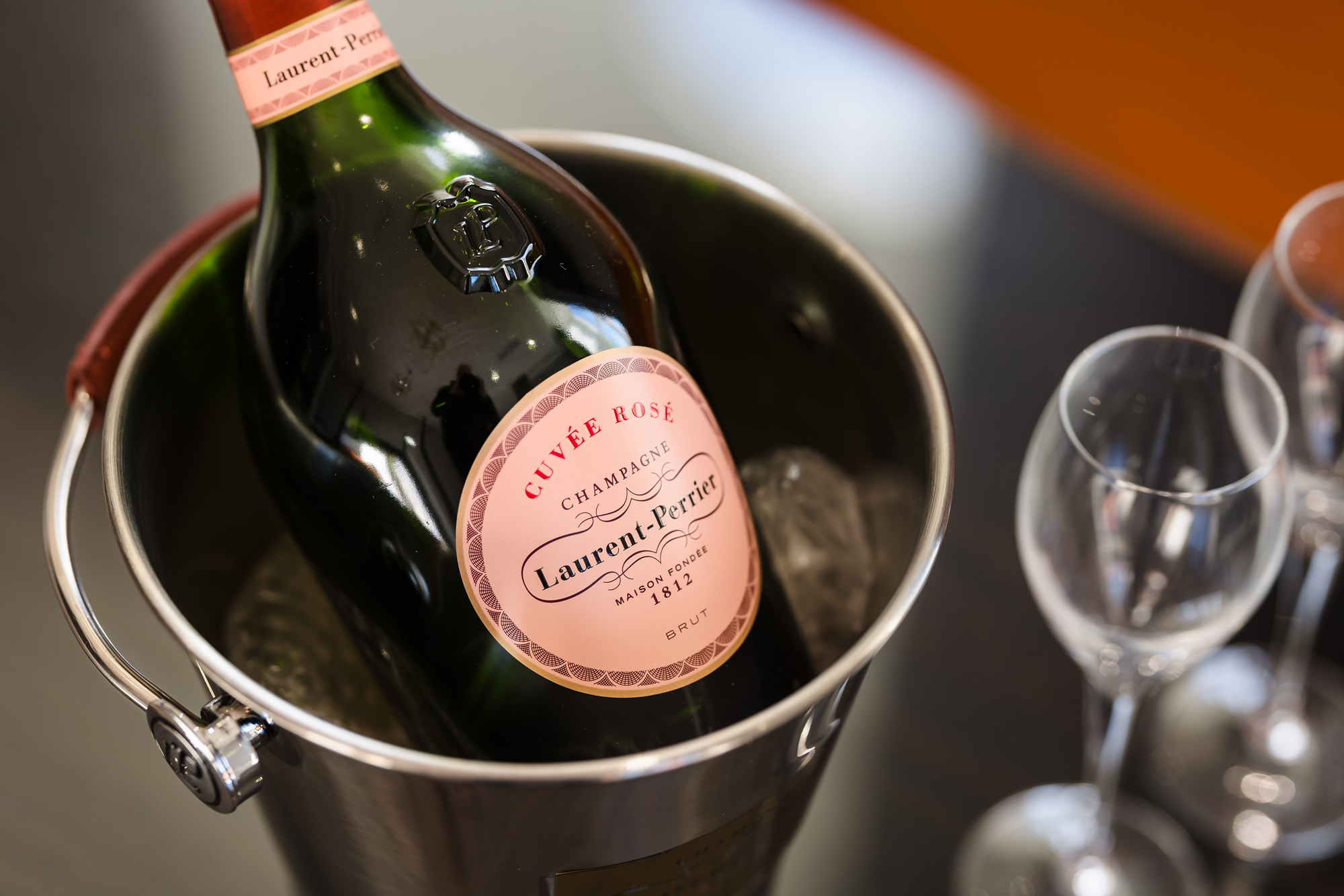 A bottle of Laurent-Perrier Cuvée Rosé Champagne rests in an ice bucket beside two empty wine glasses on a table.