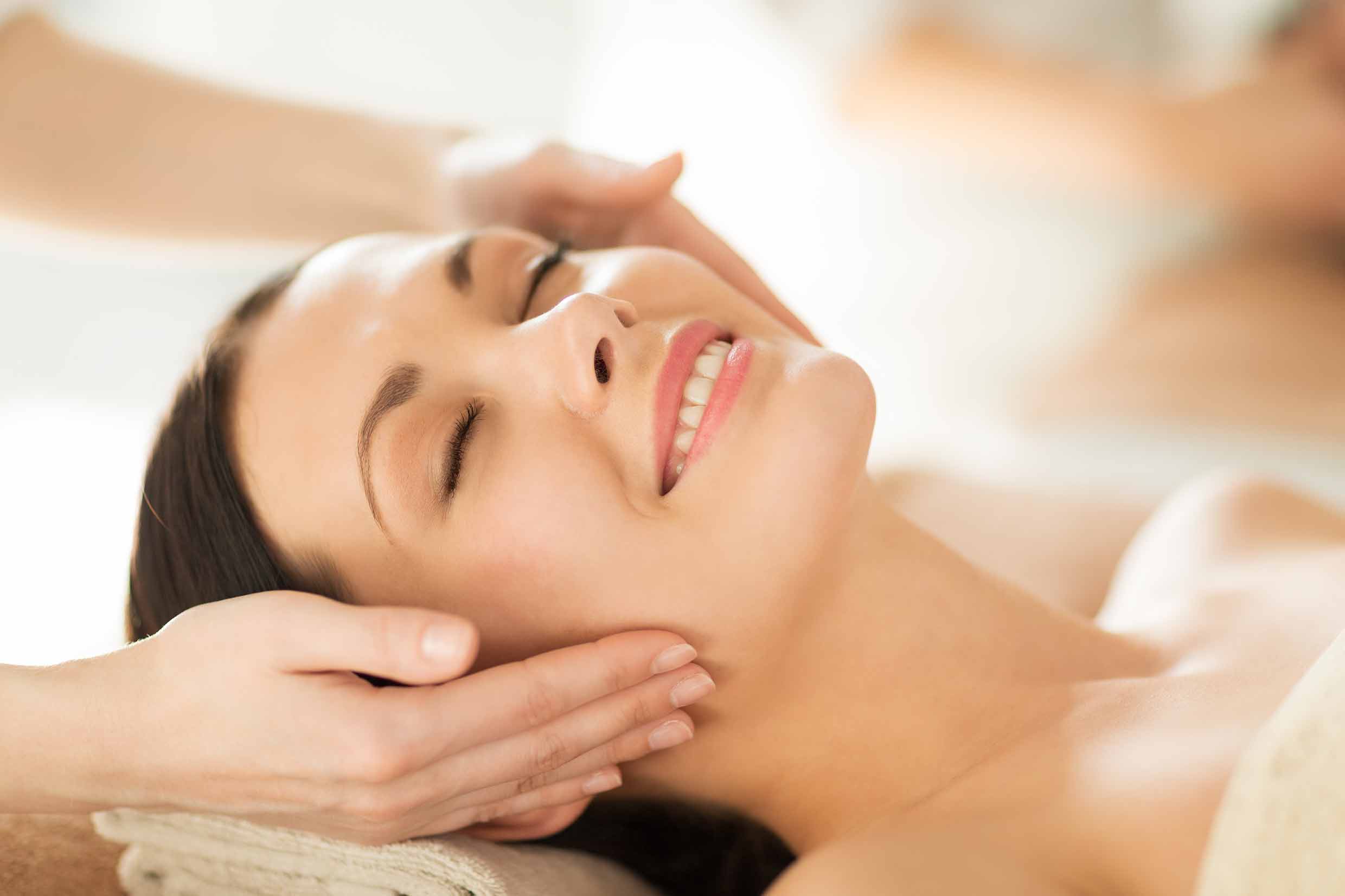 A person with closed eyes smiles while receiving a facial massage, hands gently holding their head, lying on a cushioned surface.