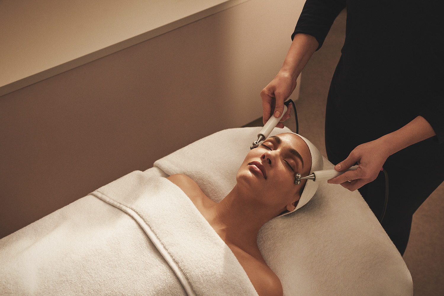 Person receiving a facial treatment with tools on a spa bed, covered with a white towel, and wearing a headband.